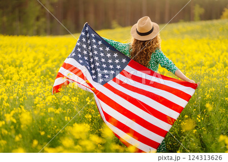 Young woman with american flag on blooming meadow. Independence Day. USA flag fluttering in the wind 124313626