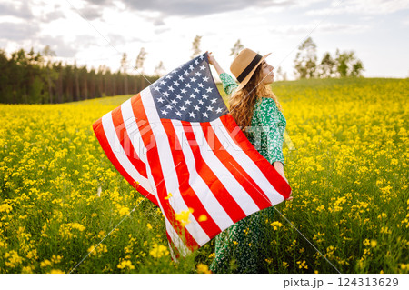 Young woman with american flag on blooming meadow. Independence Day. USA flag fluttering in the wind Young woman with american flag on blooming meadow. Independence Day. USA flag fluttering in the wind 124313629