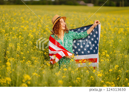 Young woman with american flag on blooming meadow. Independence Day. USA flag fluttering in the wind Young woman with american flag on blooming meadow. Independence Day. USA flag fluttering in the wind 124313630