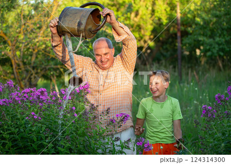 A man waters flowers with a watering can while his grandson watches and laughs. The scene is peaceful and serene, the two men enjoying the simple pleasure of tending to a garden. 124314300