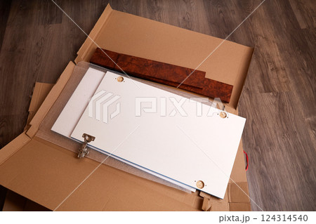 Cabinet components are arranged on the floor, showcasing a white door, a brown panel, and packaging materials.  Cabinet components are arranged on the floor, showcasing a white door, a brown panel, and packaging materials.  124314540