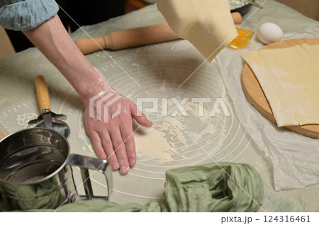 Woman's hand holding a sheet of dough and sprinkles flour worktop. Rolling pin, cutter, eggs, green towel on kitchen table. Home cooking baking puffs 124316461