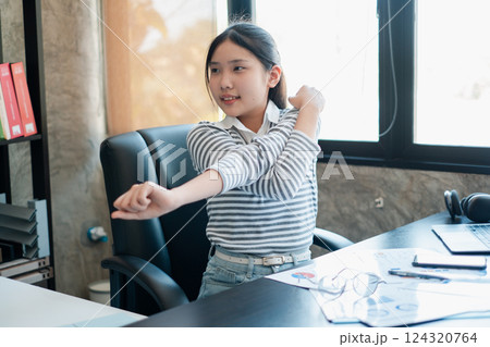 A young girl takes a break to stretch at her office desk, promoting a healthy lifestyle in a modern workspace. 124320764