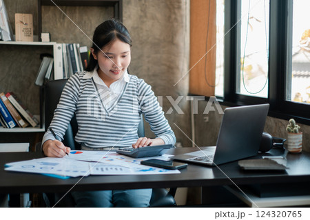 A young woman working at a desk, analyzing financial documents with a laptop in a modern office setting. A young woman working at a desk, analyzing financial documents with a laptop in a modern office setting. 124320765