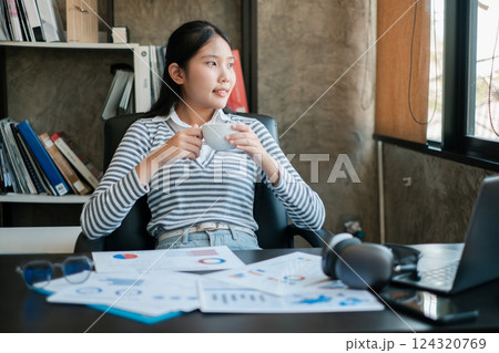 Young woman in striped shirt taking a coffee break in a modern office setting with documents and a laptop. Young woman in striped shirt taking a coffee break in a modern office setting with documents and a laptop. 124320769