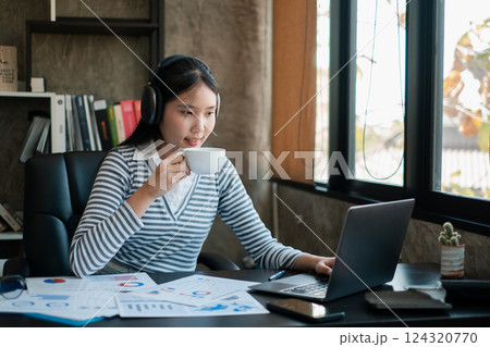Young woman in headphones working on a laptop, analyzing business reports, and enjoying coffee in a modern office. Young woman in headphones working on a laptop, analyzing business reports, and enjoying coffee in a modern office. 124320770