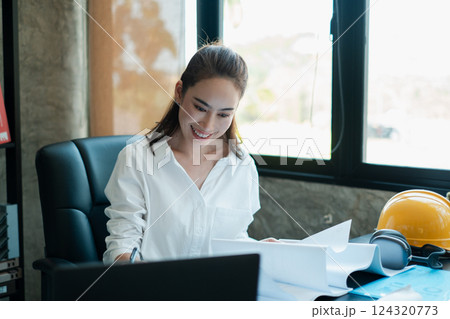 Smiling woman in a modern office setting, reviewing architectural plans with safety gear and headphones on the desk. Smiling woman in a modern office setting, reviewing architectural plans with safety gear and headphones on the desk. 124320773