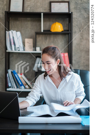 Smiling woman in white shirt working on a laptop with documents in a contemporary office environment. Smiling woman in white shirt working on a laptop with documents in a contemporary office environment. 124320774