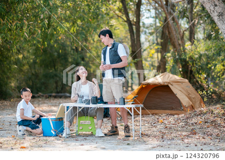 A family of three enjoys a peaceful camping trip in the woods, sitting by a picnic table with a tent in the background. 124320796