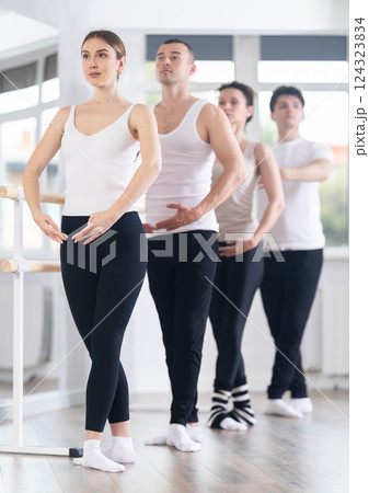 Cheerful slim active girl training various ballet poses during group classes with ballet dancers at barre in choreography studio Cheerful slim active girl training various ballet poses during group classes with ballet dancers at barre in choreography studio 124323834