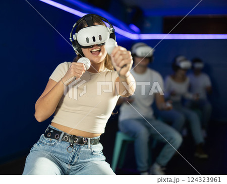 In darkened hall of gaming club, girl in VR helmet takes part in group game In darkened hall of gaming club, girl in VR helmet takes part in group game 124323961