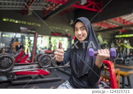 Arab middle eastern muslim woman working out while lifting a dumbbell with thumb up gesture at the gym. Fitness exercise and healthy lifestyle concept 124325721