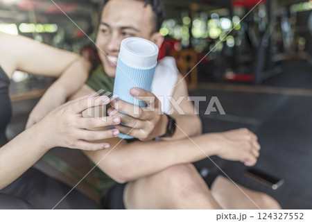 Indonesian southeast asian personal trainer man giving sporty plastic water bottle to chinese east asian woman at the gym. Fitness exercise and healthy lifestyle concept 124327552