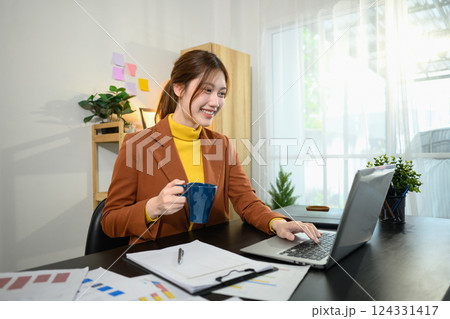 Confident young businesswoman holding a coffee mug and using laptop in a stylish home office Confident young businesswoman holding a coffee mug and using laptop in a stylish home office 124331417