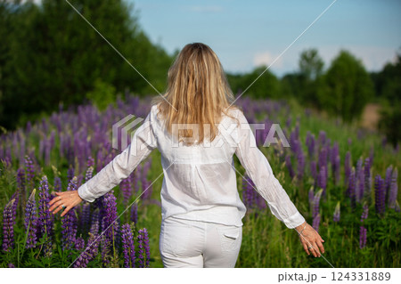 A woman walks with her back to the camera through a field of purple lupine flowers. She is dressed in white and enjoys the beauty of the flowers. A woman walks with her back to the camera through a field of purple lupine flowers. She is dressed in white and enjoys the beauty of the flowers. 124331889