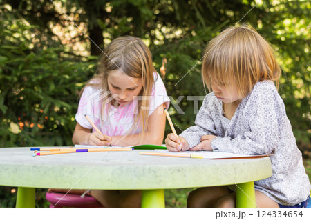 Two children sketching with colorful pens at a table in a green outdoor setting Two children sketching with colorful pens at a table in a green outdoor setting 124334654