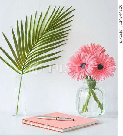serene summer workspace with pink flowers in glass vase, palm leaf in another vase, and pink notebook with pen on white desk 124344220
