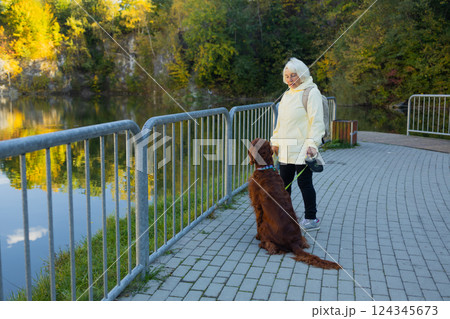 Happy senior 60s woman crossbreed looking out over a lake with her best friend irish setter dog wear casual clothes in the autumn forest.  124345673