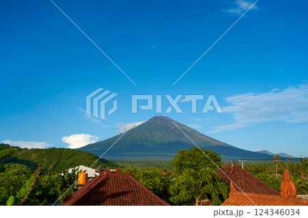 Panorama of Mount Agung and rice fields on the island of Bali. View of the mountain against a background of palm trees and a cornfield. Panorama of Agung volcano covered with clouds on a sunny day. Panorama of Mount Agung and rice fields on the island of Bali. View of the mountain against a background of palm trees and a cornfield. Panorama of Agung volcano covered with clouds on a sunny day. 124346034