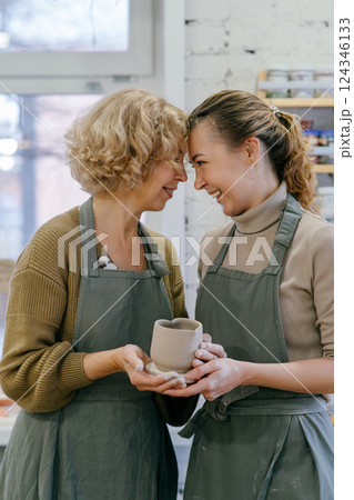 Elderly woman and young artisan holding handmade ceramic cup in pottery studio Family bond, mother and daughter enjoying creative success in pottery making Elderly woman and young artisan holding handmade ceramic cup in pottery studio Family bond, mother and daughter enjoying creative success in pottery making 124346133