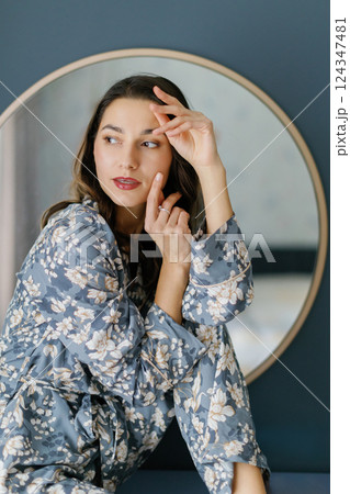 Woman wearing pajamas sitting on furniture in bedroom with round mirror and wall lamps Woman wearing pajamas sitting on furniture in bedroom with round mirror and wall lamps 124347481