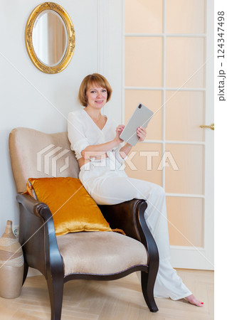 Smiling mature woman holding tablet, relaxing on a armchair at home in the living room. 124347498