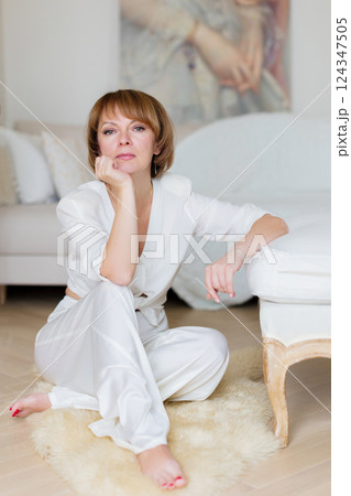 Happy adult woman in white casual clothes sitting on floor near couch spending time in living room at home. Rest relax good mood leisure lifestyle concept. 124347505