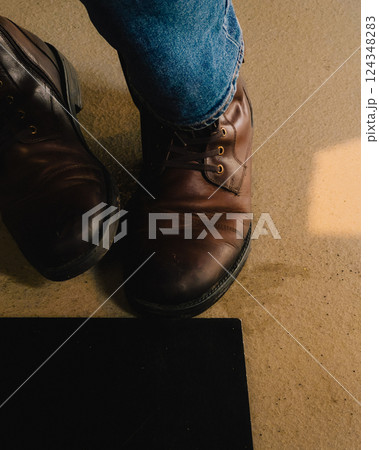 Close up of brown leather boots worn by man in blue jeans, standing on beige floor illuminated by warm, natural light. Man wearing brown leather boots and blue jeans Close up of brown leather boots worn by man in blue jeans, standing on beige floor illuminated by warm, natural light. Man wearing brown leather boots and blue jeans 124348283