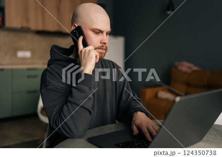 Serious freelancer male working from remote home office, talking on mobile phone and using laptop computer, managing business and multitasking in home environment. Concept of remote job, technology. 124350738