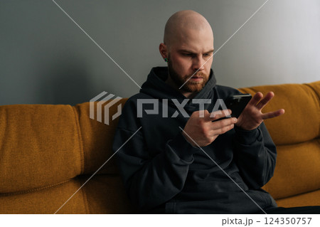 Close-up of serious young bald man with beard and hoodie using smartphone while sitting on comfortable sofa, browsing internet, social media or mobile application. Concept of modern technology. Close-up of serious young bald man with beard and hoodie using smartphone while sitting on comfortable sofa, browsing internet, social media or mobile application. Concept of modern technology. 124350757