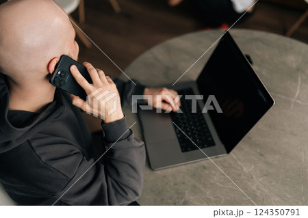 High-angle view of bald freelancer male working remotely, communicating on smartphone and typing on laptop, managing business while multitasking in contemporary home workspace, sitting at desk. High-angle view of bald freelancer male working remotely, communicating on smartphone and typing on laptop, managing business while multitasking in contemporary home workspace, sitting at desk. 124350791