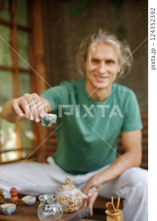 Blonde man preparing for a tea ceremony. Man sitting in outdoor Japanese spring garden in backyard porch of zen home. Focus on a cup in man`s hands. Blonde man preparing for a tea ceremony. Man sitting in outdoor Japanese spring garden in backyard porch of zen home. Focus on a cup in man`s hands. 124352392