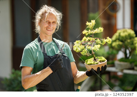 Blonde caucasian man planting flowers in pot with garden tools. Man wearing green t-shirt and black apron. Man spending peaceful morning in garden hear house. 124352397
