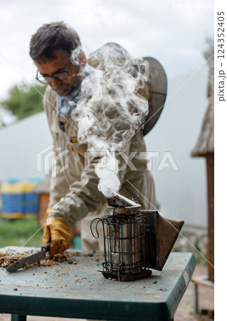 Experienced senior apiarist in his apiary setting a fire in a bee smoker. Beekeeper harvesting honey. Bee smoker is used to calm bees before frame removal. Experienced senior apiarist in his apiary setting a fire in a bee smoker. Beekeeper harvesting honey. Bee smoker is used to calm bees before frame removal. 124352405