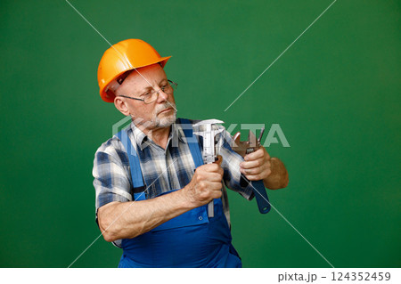 Studio portrait of a man isolated on green background. Seniour man worker in a blue plaid shirt and blue overall holds a straight pipe wrench or spanner. Man wearing eyeglasses and orange helmet. Studio portrait of a man isolated on green background. Seniour man worker in a blue plaid shirt and blue overall holds a straight pipe wrench or spanner. Man wearing eyeglasses and orange helmet. 124352459