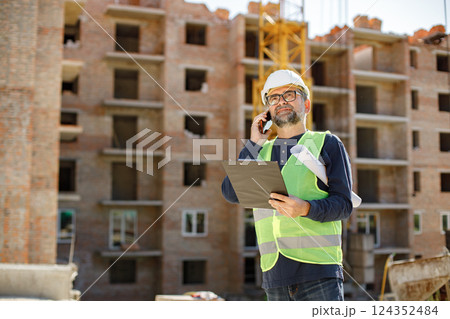 Construction site manager standing wearing safety vest and helmet, thinking at construction site. Architect with papers at a construction site. Man talking on the phone. Construction site manager standing wearing safety vest and helmet, thinking at construction site. Architect with papers at a construction site. Man talking on the phone. 124352484