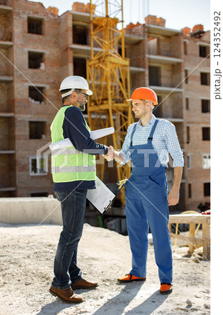 Worker and architect watching some details on a construction. Two engineers talking at building site with construction structure in background. Men wearing helmets. Worker and architect watching some details on a construction. Two engineers talking at building site with construction structure in background. Men wearing helmets. 124352492