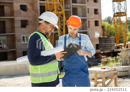 Worker and architect watching some details on a construction. Two engineers talking at building site with construction structure in background. Men wearing helmets. 124352511
