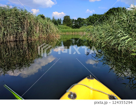 Man floating on inflatable boat along small river between reeds. Person floating on yellow inflatable boat on mirror surface of water of small pond with reflection of blue sky on calm summer sunny day Man floating on inflatable boat along small river between reeds. Person floating on yellow inflatable boat on mirror surface of water of small pond with reflection of blue sky on calm summer sunny day 124355294