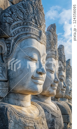 Stone statues of serene faces in traditional Khmer style, lined up under blue sky 124356095