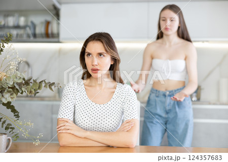 Angry mother sitting at the kitchen table, daughter standing behind Angry mother sitting at the kitchen table, daughter standing behind 124357683