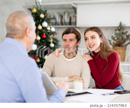 Male realtor discussing home choice with couple in real estate agency office, offering various options for residence against the background of Christmas tree Male realtor discussing home choice with couple in real estate agency office, offering various options for residence against the background of Christmas tree 124357963