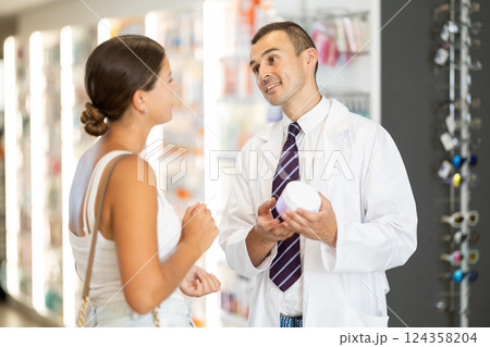 Male consultant pharmacist holds box in hands and offers girl client to purchase remedy Male consultant pharmacist holds box in hands and offers girl client to purchase remedy 124358204