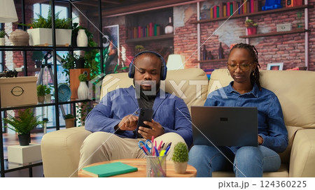 Young woman working on freelance tasks while her partner browses phone, modern couple using their devices sitting on the couch in the living room. People enjoying time at home. Camera A. Young woman working on freelance tasks while her partner browses phone, modern couple using their devices sitting on the couch in the living room. People enjoying time at home. Camera A. 124360225