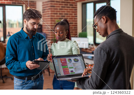 Colleagues utilizing digital technology during their discussion in modern workspace. Black man using laptop to retrieve marketing data as his male and female coworkers review strategy plans on tablet. 124360322