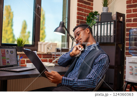 Financial advisor reviewing business analytics on clipboard and having phone discussion with client. Black businessman reading company documents and speaking to coworker on a landline call. Financial advisor reviewing business analytics on clipboard and having phone discussion with client. Black businessman reading company documents and speaking to coworker on a landline call. 124360363