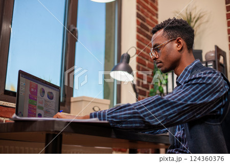 African American manager at office desk reviewing company research updates on his laptop. Male entrepreneur seated in red brick wall room, using digital device to prepare business project presentation African American manager at office desk reviewing company research updates on his laptop. Male entrepreneur seated in red brick wall room, using digital device to prepare business project presentation 124360376