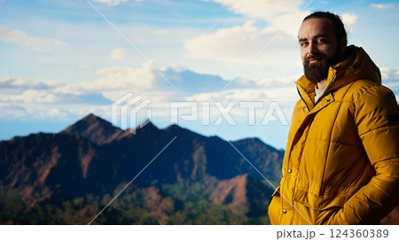 Hiker dressed in warm clothing stands on a mountain peak after climbing to the top, gazing at the breathtaking landscape of rugged hills and vast sky. Adventure with scenic view. Camera A. Hiker dressed in warm clothing stands on a mountain peak after climbing to the top, gazing at the breathtaking landscape of rugged hills and vast sky. Adventure with scenic view. Camera A. 124360389