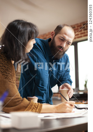 Bearded man holding coffee cup and providing input during business meeting with female analyst at office desk. Closeup on caucasian coworkers having warm beverage and reviewing financial paperwork. Bearded man holding coffee cup and providing input during business meeting with female analyst at office desk. Closeup on caucasian coworkers having warm beverage and reviewing financial paperwork. 124360393