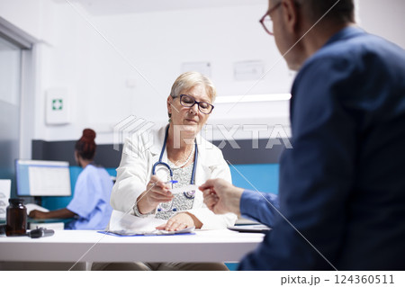 Male pensioner patient receives medical advice in form of written document from retired doctor in clinic office. Elderly female physician in lab coat handing over prescription to senior man. 124360511